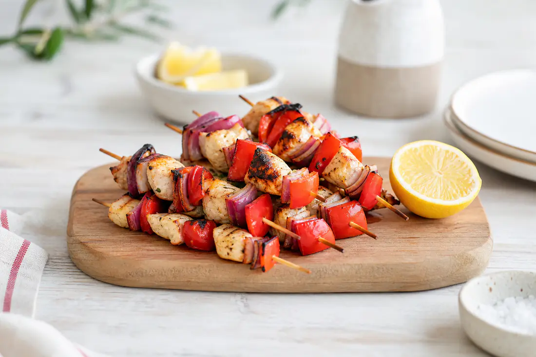 Spiedini di pollo con peperoni e cipolla rossa, dorati e leggermente bruciacchiati, serviti su un tagliere di legno con limone