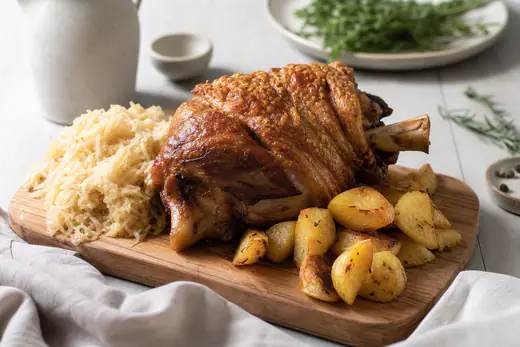 Stinco di maiale intero con la pelle dorata e croccante, servito con patate arrosto e crauti su un tagliere con un coltello