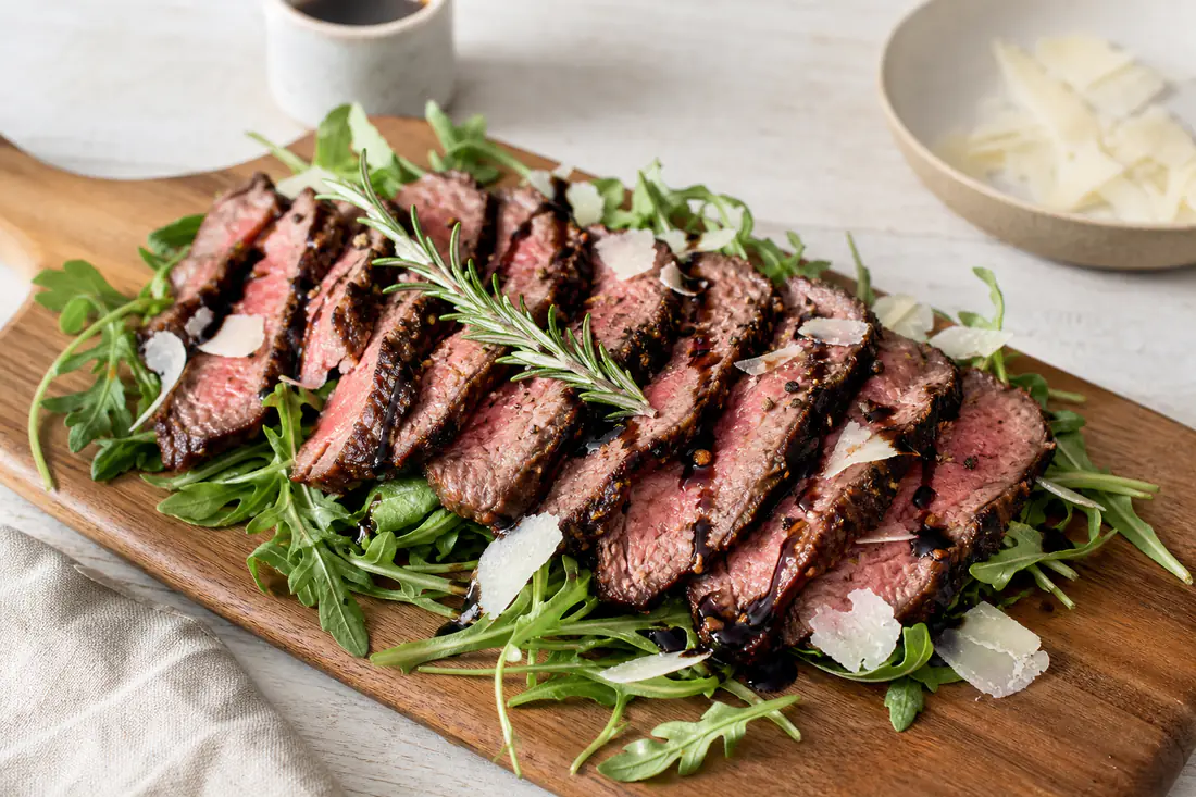 Tagliata di manzo a fette rosate su un letto di rucola con scaglie di grana, un filo di aceto balsamico e un rametto di rosmarino
