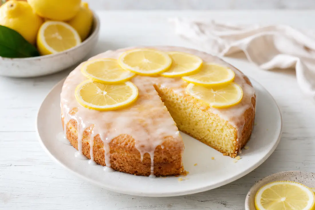 Torta al limone rotonda con glassa bianca leggera e fettine di limone, una fetta tagliata che mostra la mollica soffice e gialla