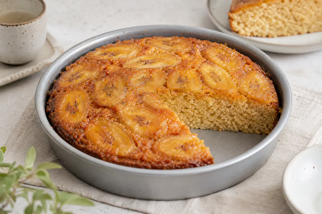 Torta alla banana rotonda in una tortiera tonda, con fette di banana dorate sulla superficie e una fetta tagliata dalla mollica umida