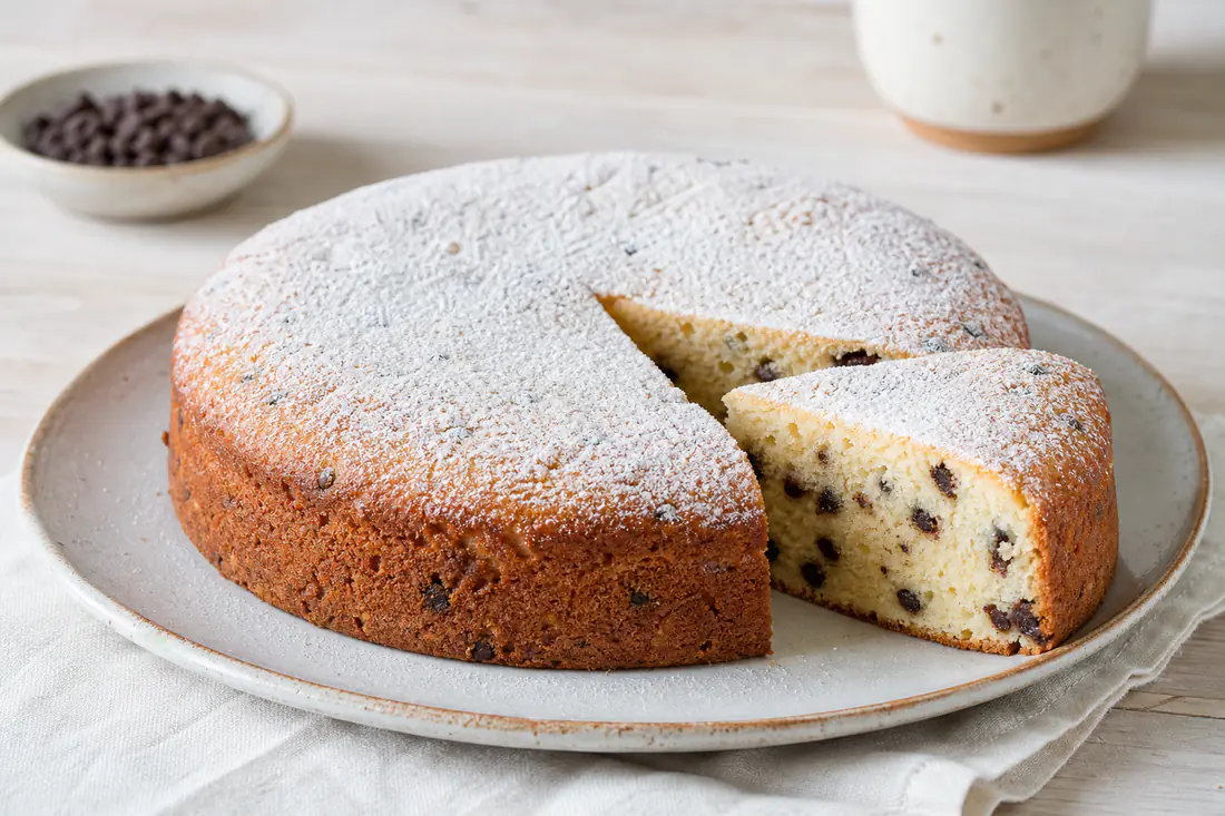 Torta rotonda di ricotta dorata con zucchero a velo, una fetta tagliata che mostra la mollica chiara con gocce di cioccolato