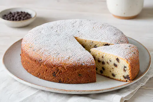 Torta rotonda di ricotta dorata con zucchero a velo, una fetta tagliata che mostra la mollica chiara con gocce di cioccolato