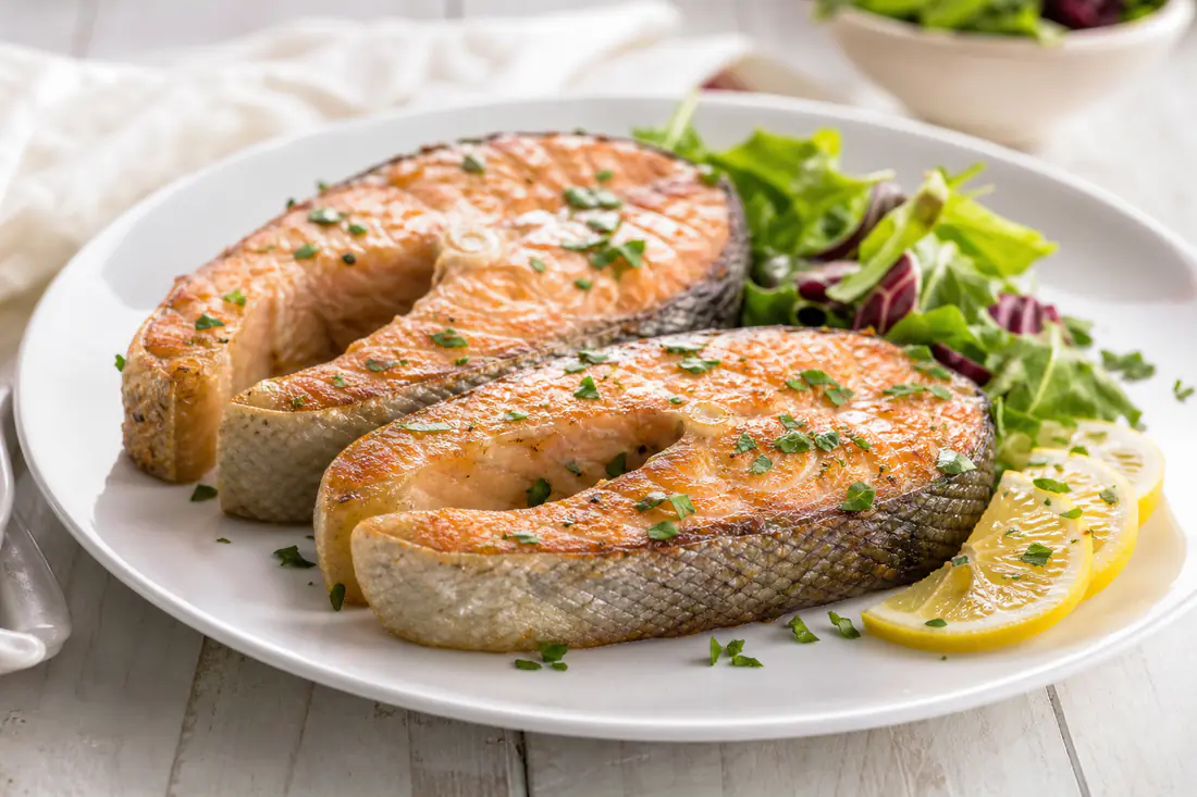 Due tranci di salmone con osso centrale e pelle dorata, serviti su un piatto con fette di limone, prezzemolo e un contorno di insalata