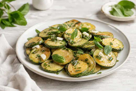 Rondelle di zucchine dorate marinate alla scapece con foglie di menta fresca e spicchi d’aglio, in un piatto di ceramica bianca